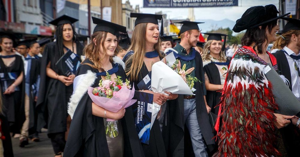 Massey graduates in academic gowns and caps process along a crowded street, one carrying flowers.