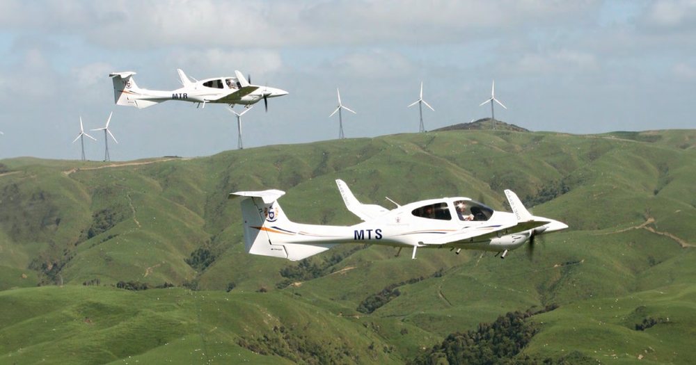 Two light aircraft flying in formation over green hill country dotted with wind turbines.