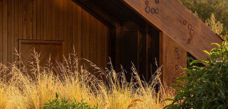 A timber-clad building with carved circular patterns along its beam, framed by golden tussock grass.